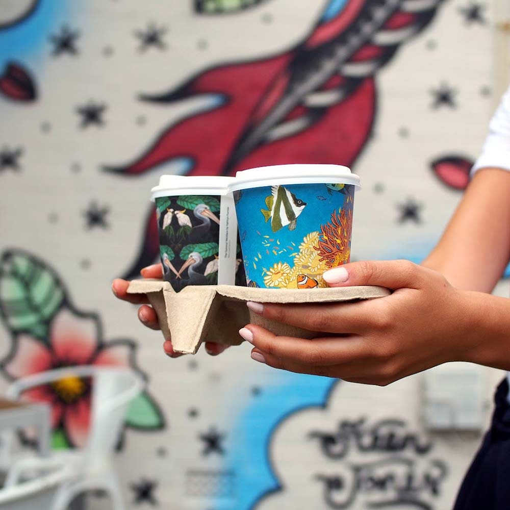 Person holding a Pulp BioCup 2-cup tray with two colorful coffee cups in front of a mural featuring flowers and abstract shapes.
