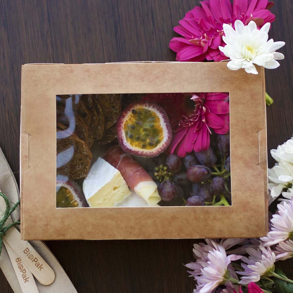 Cardboard BioBoard Lunch Box with a clear window reveals cheese, grapes, crackers, prosciutto, and passion fruit. Arranged beside pink and white flowers and wooden cutlery for a charming presentation.
