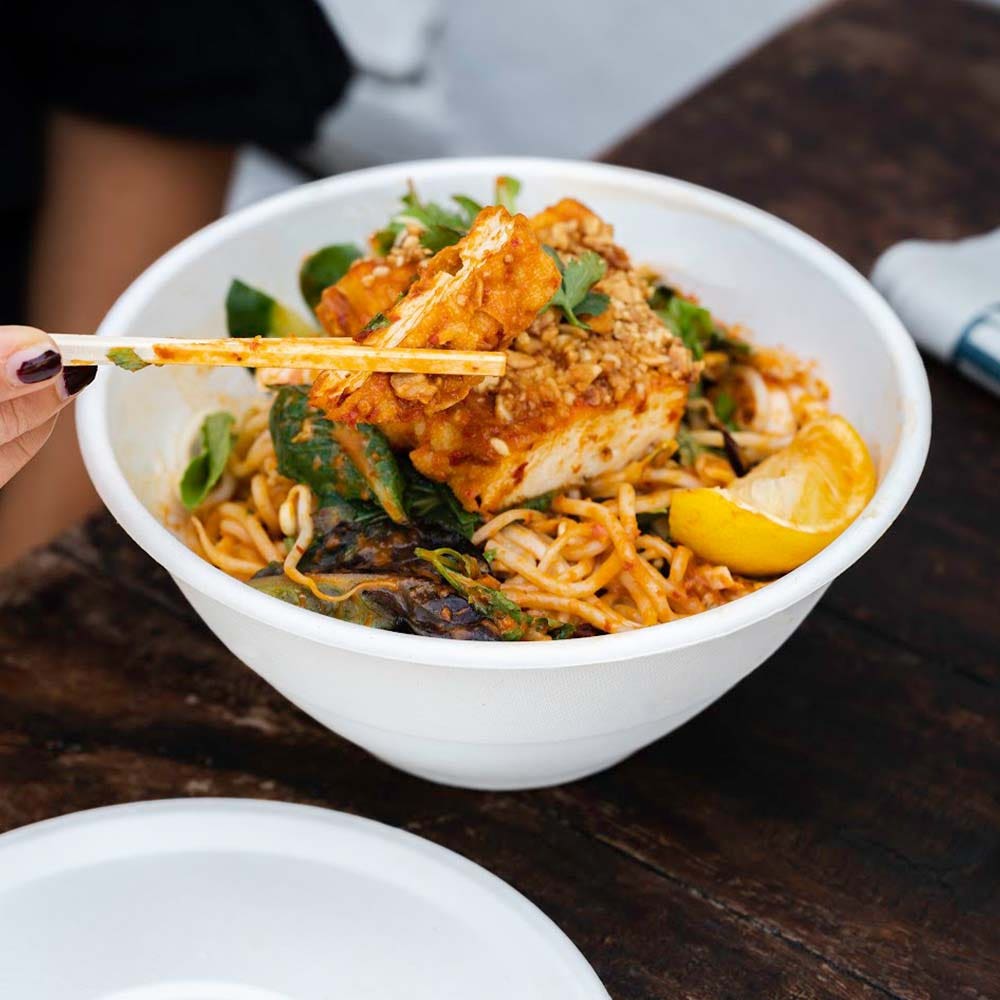 A hand using chopsticks picks up noodles from a 420ml BioBowl filled with tofu, vegetables, sauce, and a lemon wedge on a dark wooden table.