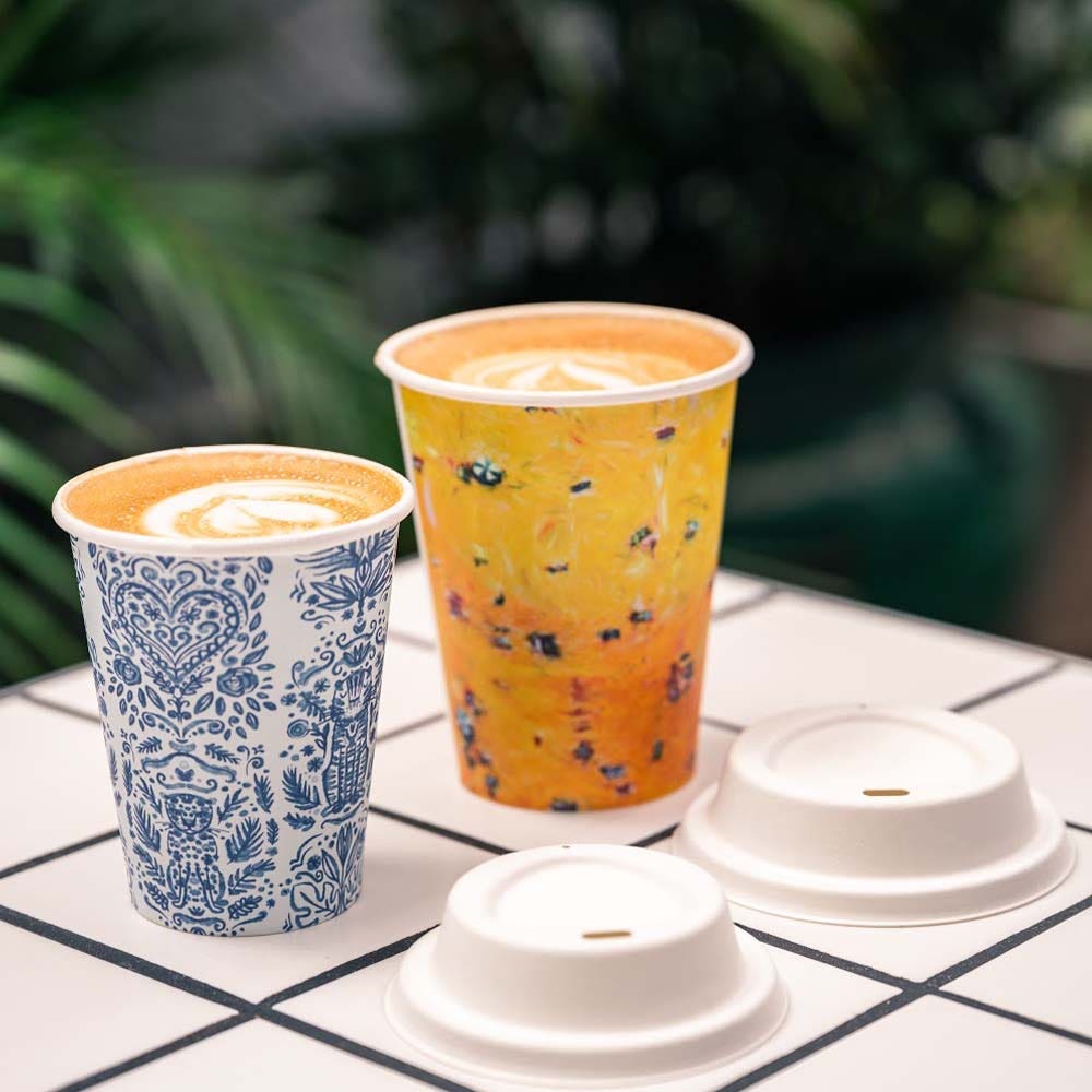 Two cups of coffee with latte art in colorful Art Series BioCup single wall cups sit on a tiled surface next to two white cup lids, with greenery blurred in the background.
