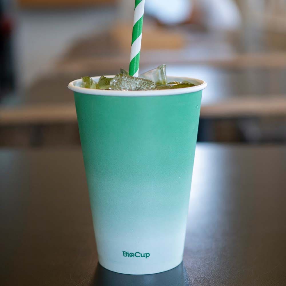 A green and white Green Cold Paper BioCup with ice and a green striped paper straw sits on a dark surface.