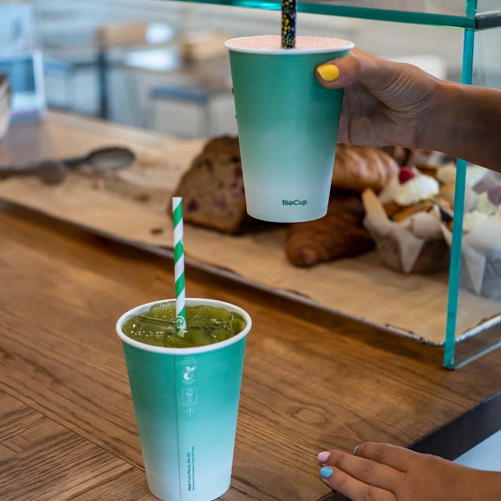 Two hands holding 300ml paper cups with striped straws, one filled with a green drink and the other with a pink drink, rest on a wooden counter near pastries.