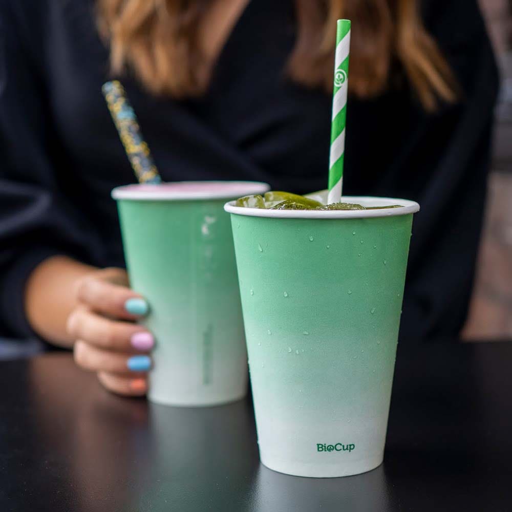 Person holding a Green Cold Paper BioCup with a striped paper straw; another 8oz Paper Cup sits on the table, both labeled "BioCup.
