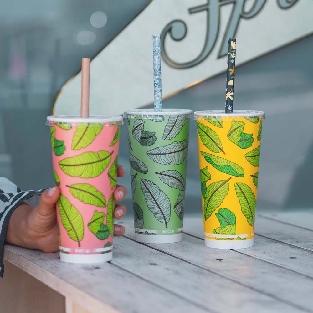 Three colorful 650ml cold paper cups with leaf patterns and patterned straws rest on a wooden table; a hand holds the pink bio cup on the left.