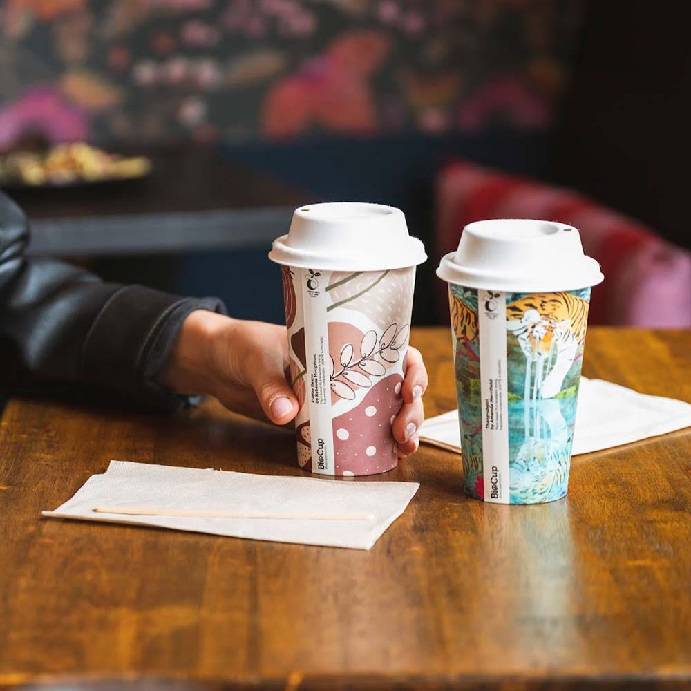 Two decorative 12oz BioCup takeaway coffee cups with lids sit on a wooden table; a hand holds one cup. Two napkins are placed beside the cups.