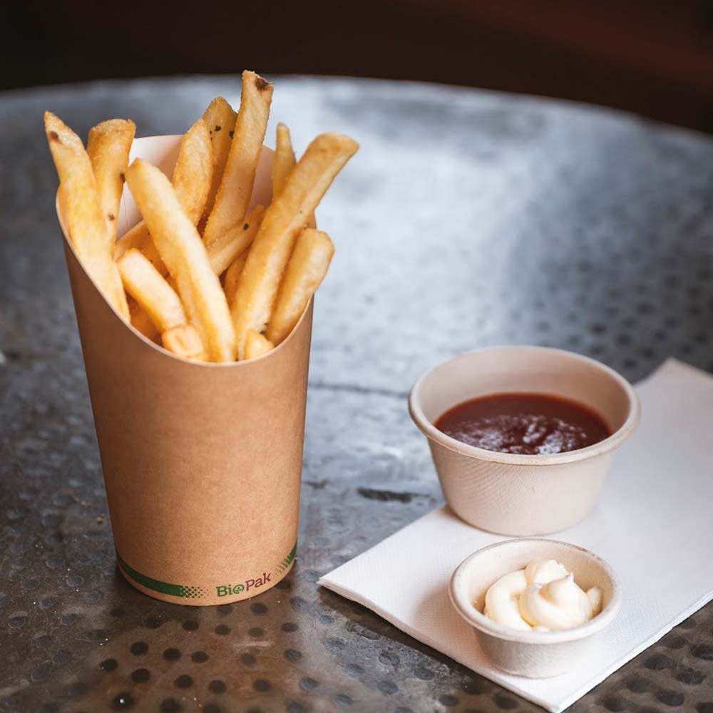 A cardboard container of French fries sits on a metal table next to small cups of ketchup and mayonnaise served in an eco-friendly Medium BioCup on a white napkin.