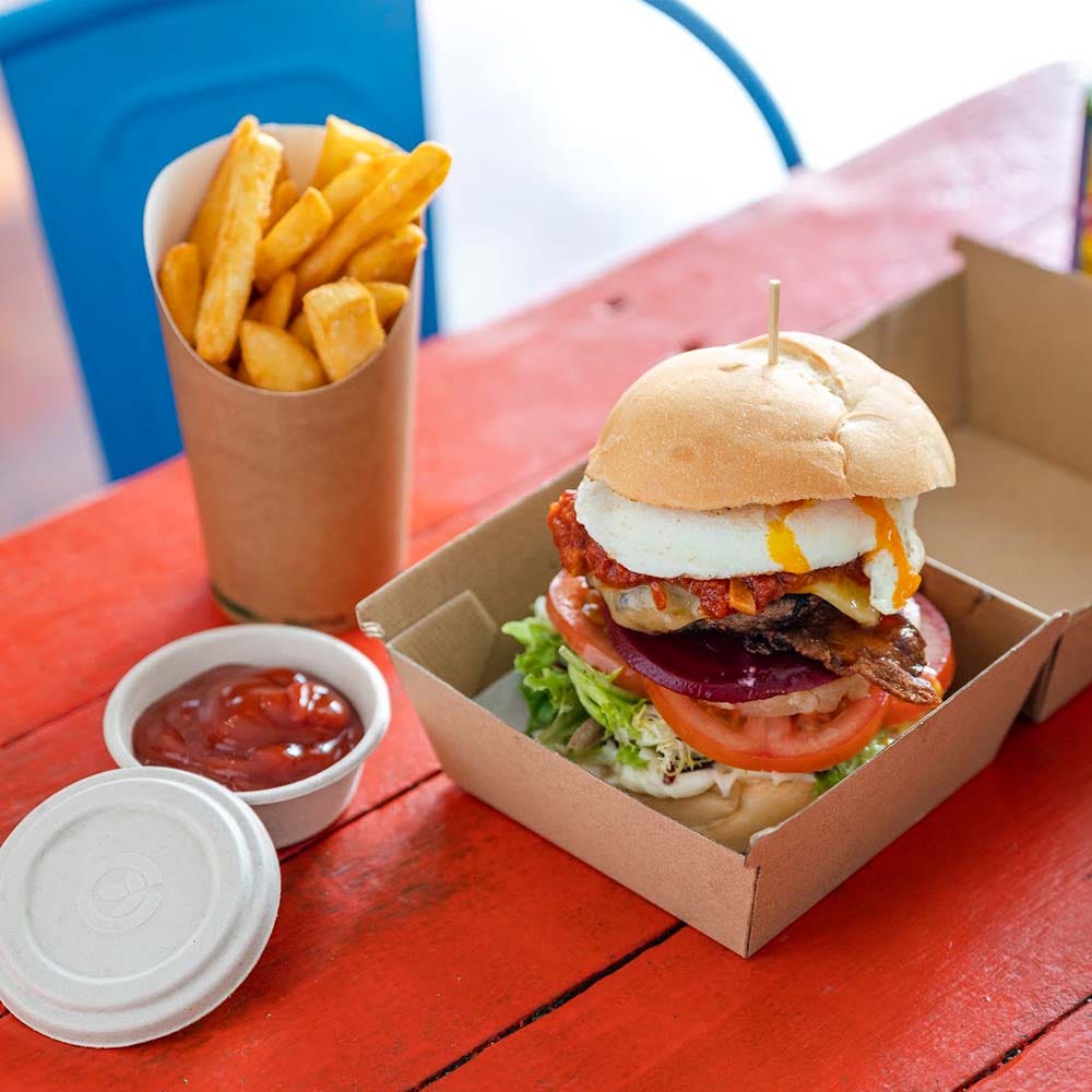 Burger with egg, bacon, lettuce, tomato, and onion in a BioBoard Box takeout container, served with a cup of fries and a container of ketchup on a red table.