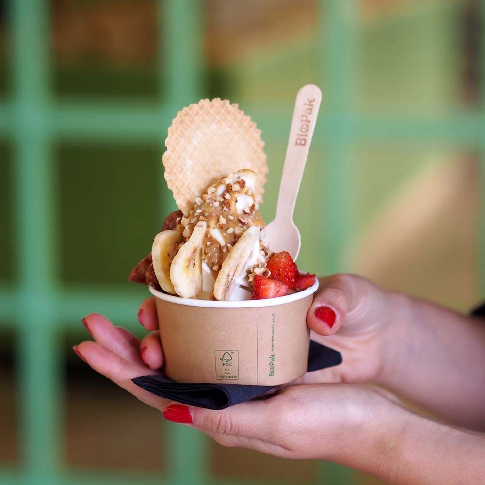 A person holds a 90ml Ice Cream Cup filled with frozen yogurt, topped with banana slices, strawberries, chopped nuts, a wafer, and a wooden spoon.