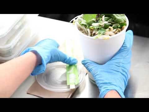 A person wearing blue gloves packs a takeout container of food, places chopsticks on the lid, and sets a biodegradable cup next to disposable containers.