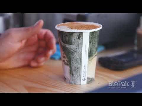 A hand reaches toward a 230ml Art Series BioCup Single Wall coffee cup with a printed design, placed on a wooden surface next to a smartphone. The cup is branded with the BioPak logo.