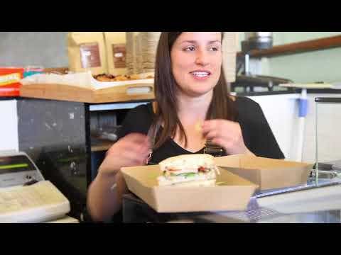 A woman sits at a counter with an open BioBoard Noodle Box containing a sandwich in front of her, gesturing with her hands.