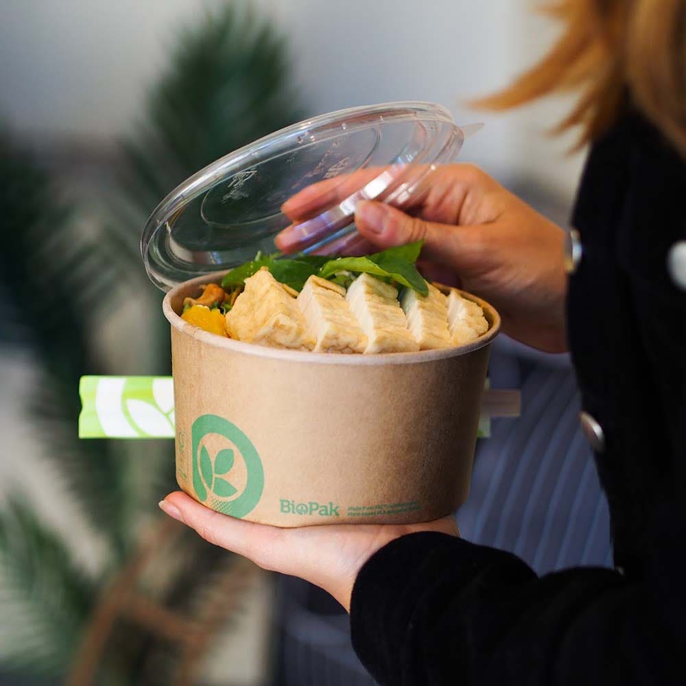 A person holds a Large Kraft BioBowl with a clear plastic lid, containing sliced tofu, greens, and vegetables.